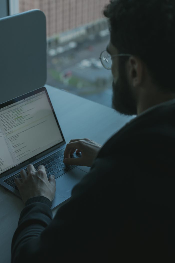 A man coding on his laptop by a window in an office setting, showcasing technology work.